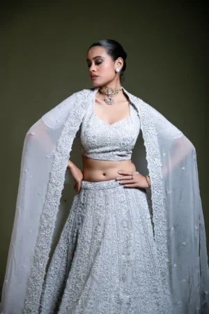 Three-quarter front view of a model posing with one hand on her hip, gazing downward, wearing a light grey heavily embroidered organza lehenga set. The image highlights the dense tonal floral threadwork, beads, and sequins on the choli and flared skirt, along with the embroidered border of the floor-length cape dupatta. Styled with a gold polki choker necklace and gold bangle.